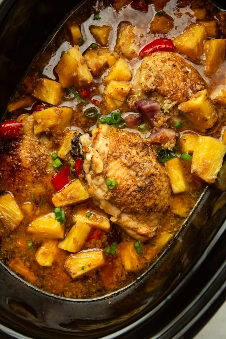 a black crockpot full of cooked chicken thighs, pineapple chunks, red bell peppers, purple onions, and chopped green onions