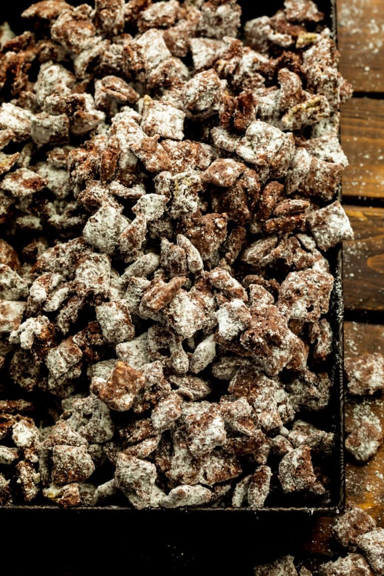 powdered sugar coated dark chocolate muddy buddies in a shallow serving tray on a wooden table