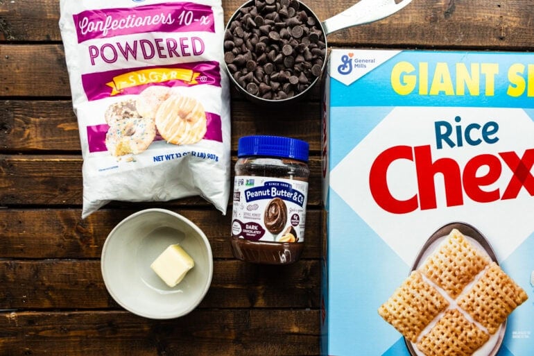 All ingredients for dark chocolate Mud Buddy including a box of Mickeyx cereal, chocolate chips, butter, powdered sugar and dark chocolate peanut butter sitting on wooden table