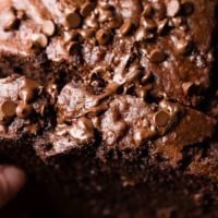 A close up photograph of a slice being picked up from a loaf of chocolate banana bread with mini chocolate chips all over the top that are starting to melt. The very best double chocolate banana bread