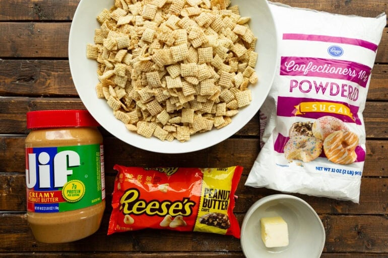 all the ingredients for peanut butter muddy buddies including a big bowl of rice chex, powdered sugar, a jar of creamy peanut butter, butter and a bag of Reese's peanut butter chips