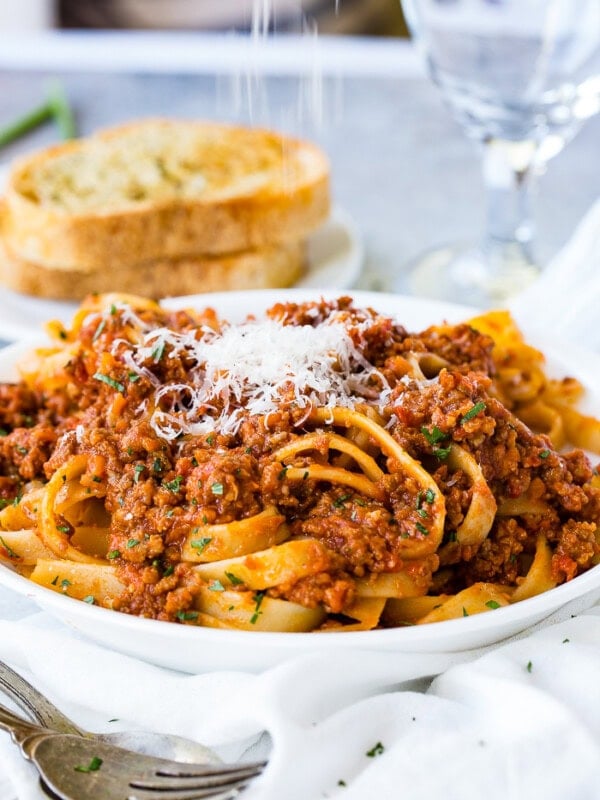 A white pasta dish full of fettuccini loaded with authentic Italian Bolognese sauce and fresh parmesan cheese on top with garlic bread in the background