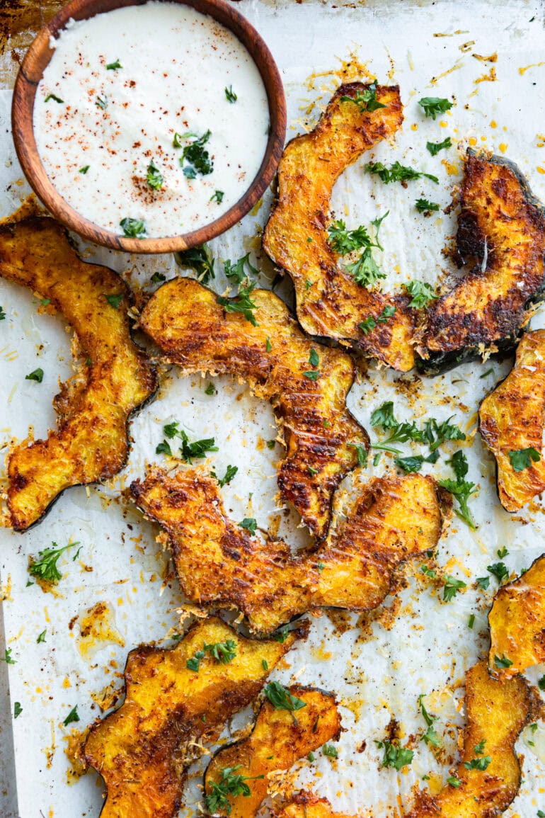 a photo of several slices of roasted acorn squash topped with golden crispy cheese and herbs with a small bowl of dip next to it