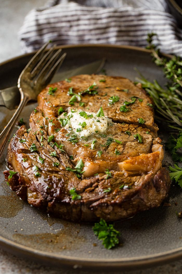 a grey plate with a medium rare ribeye steak being cut into and a fork taking a piece with garlic butter melting on top with parsley