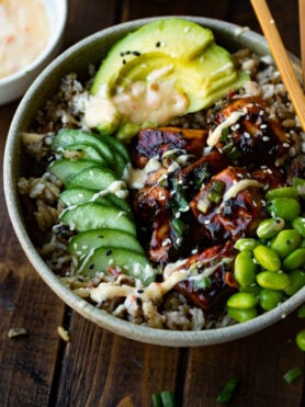 a photo of a large bowl full of cooked wild rice topped with chunks of teriyaki glazed salmon, edamame beans, sliced cucumber, sliced avocado and all topped with sprinkled sesame seeds.