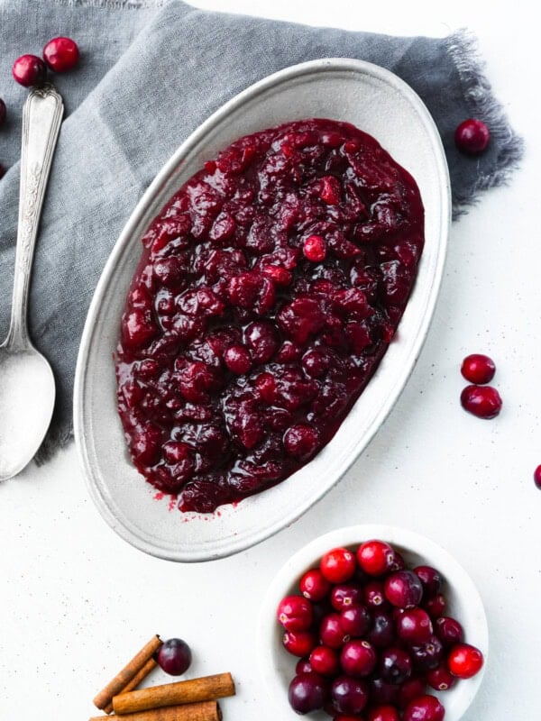 a photo of a large white serving dish full of cranberry sauce with a silver serving spoon sitting next to the bowl and a small bowl of fresh cranberries next to it.