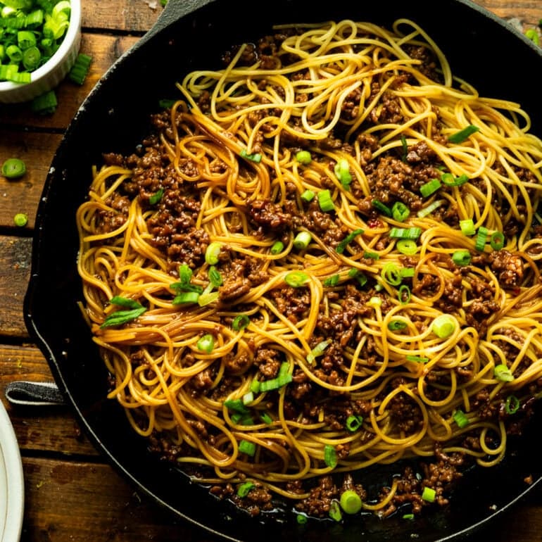 A photo of a cast iron skillet filled with cooked spaghetti mixed with ground beef and chopped green onions