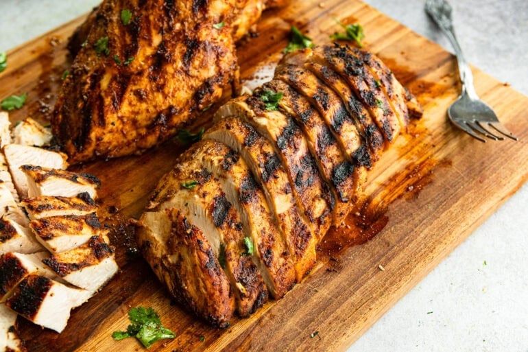 A overhead photo of seasoned grilled chicken breasts sliced thin on a wooden cutting board ready to be shredded for tacos and burrito bowls.