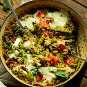 a photo of a cast iron skillet of quinoa mixed with pesto, asparagus, tomatoes and topped with melted cheese