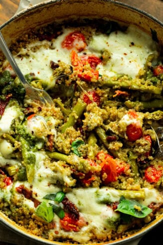 a photo of a cast iron skillet of quinoa mixed with pesto, asparagus, tomatoes and topped with melted cheese
