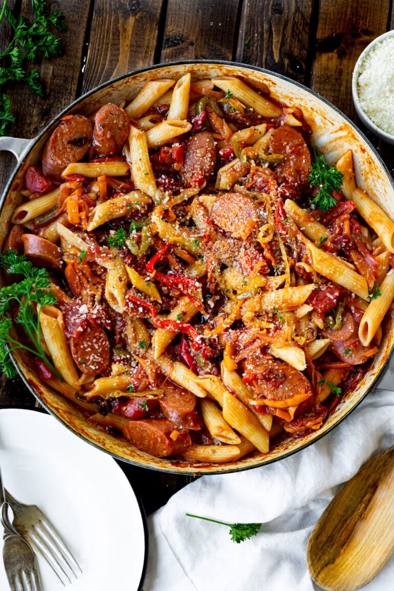 a photo of a large skillet full of slices of sausage and sauteed peppers and onions all mixed with cooked penne pasta.