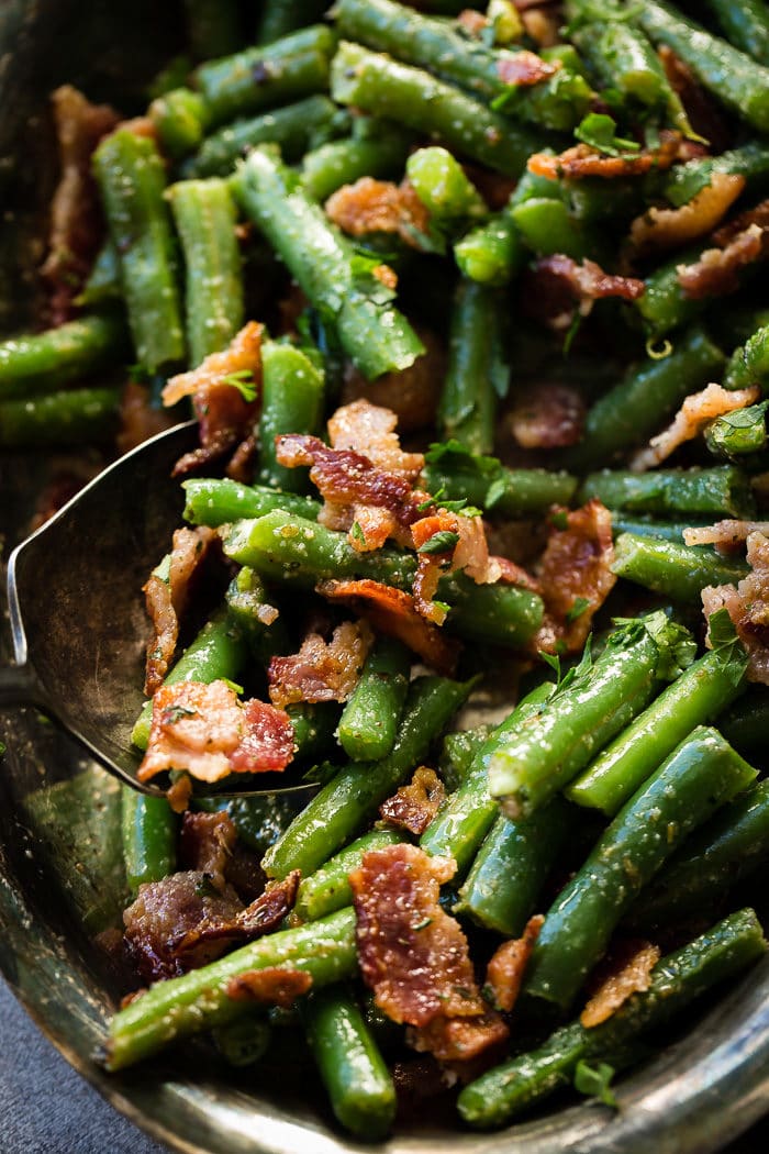 A silver serving platter and silver serving spoon dipping into a bowl of chopped green beans with crumbled bacon and a garlic seasoning