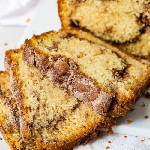 Three slices of cinnamon sugar swirled snickerdoodle bread on a plate with the rest of the loaf in the background.