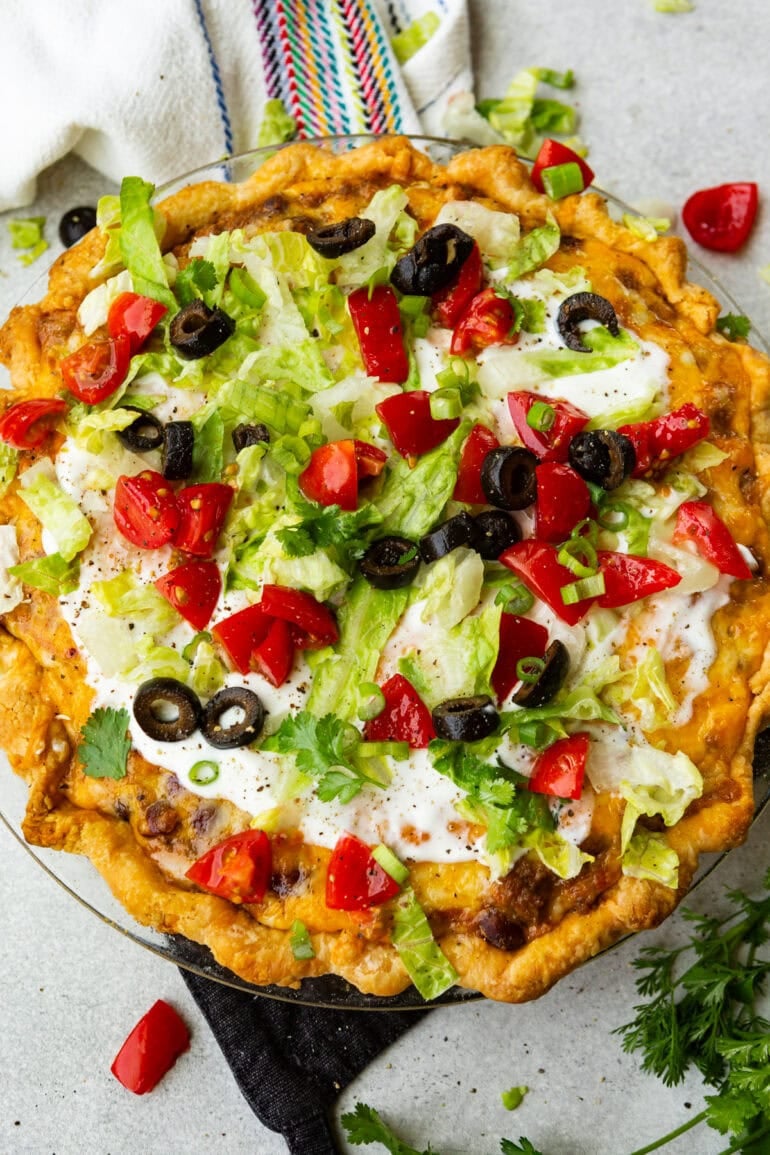 a whole taco pie with a golden crust, cheesy ground beef, topped with sour cream, shredded lettuce, diced tomatoes and black olives