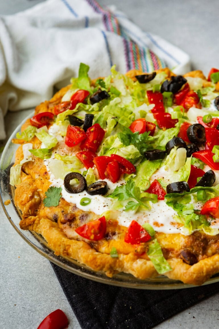a whole taco pie in a glass pie plate topped with sour cream, shredded lettuce, diced tomatoes and sliced black olives