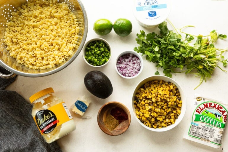 all the ingredients for mexican street corn pasta salad including corn, red onions, limes, cilantro, avocado, pasta and the dressing ingredients