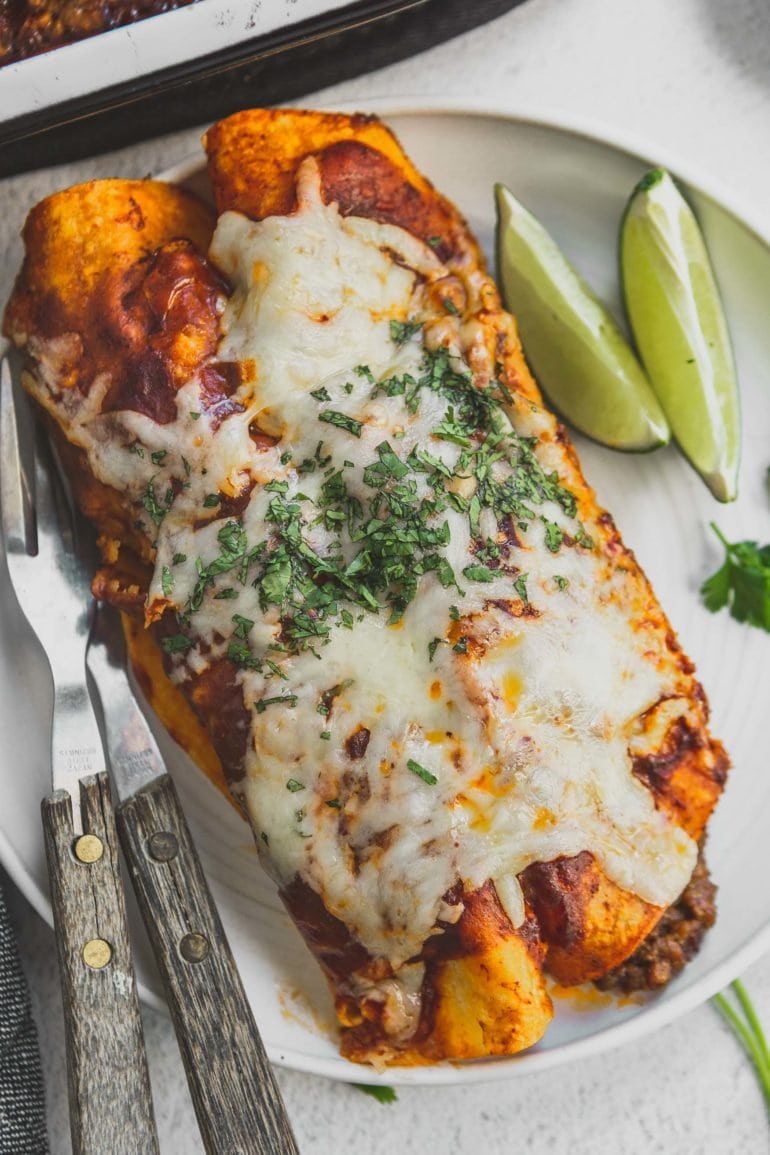 Two enchiladas on a dinner plate with lime wedges and silverware. The enchiladas are covered with melted cheese and chopped cilantro.