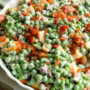 a large bowl of creamy english pea salad topped with crispy bacon bits and cubes of cheddar cheese