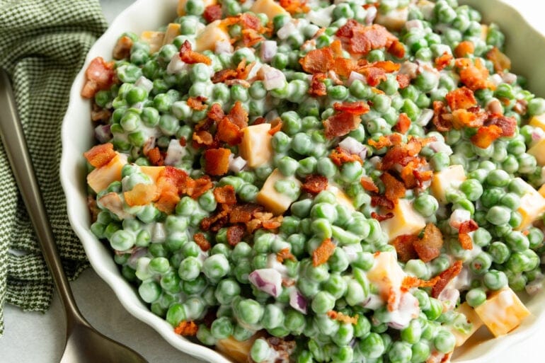 a large bowl of creamy english pea salad topped with crispy bacon bits and cubes of cheddar cheese
