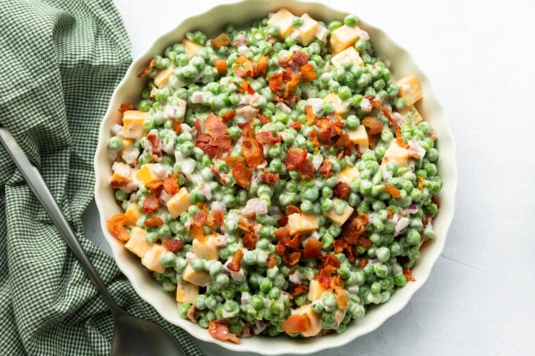 a large serving bowl full of creamy pea salad with bacon bits, red onions, and cheddar cheese