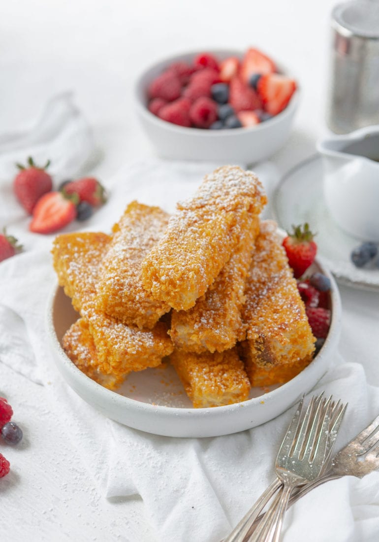 A dish of golden french toast sticks. There are sliced strawberries around the toast and in the background.