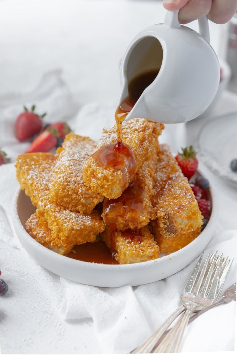 A stack of French Toast Sticks that are baked to a golden brown. they are sprinkled with powdered sugar and syrup is being poured over the top. Sliced strawberries are around the toast.