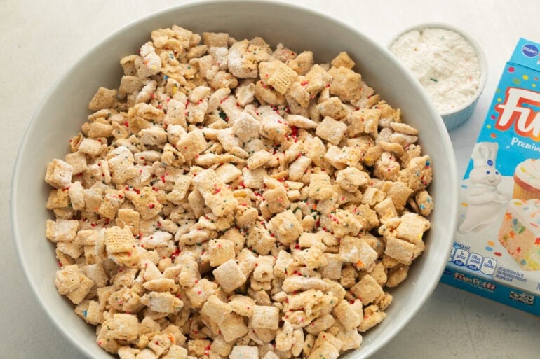 a large serving bowl of cake batter muddy buddies with a box of funfetti cake mix next to the bowl