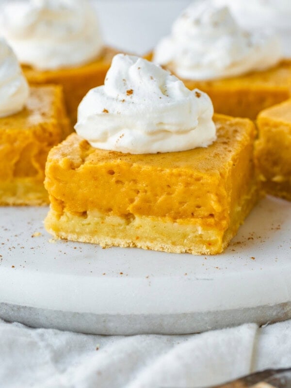 a photo of a square piece of gooey pumpkin butter cake with dollop of whipped cream on top with several pieces sitting in the background.