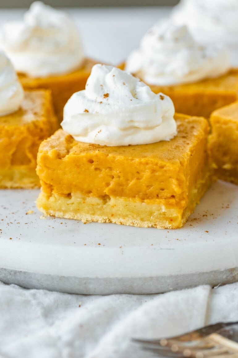 a photo of a square piece of gooey pumpkin butter cake with dollop of whipped cream on top with several pieces sitting in the background.