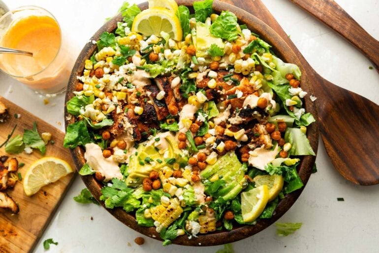 a large wooden bowl full of grilled chicken breast, sliced avocado, corn, crispy chicken peas and drizzled with southwest ranch