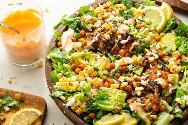 a large wooden bowl  grilled chicken salad topped with sliced avocado, crispy chickpeas and drizzled with southwest ranch