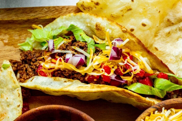 a ground beef taco topped with cheddar cheese, shredded lettuce, diced red onions and tomatoes