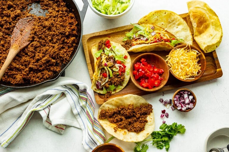 all the toppings for ground beef tacos in separate small bowls around corn tortillas filled with ground beef