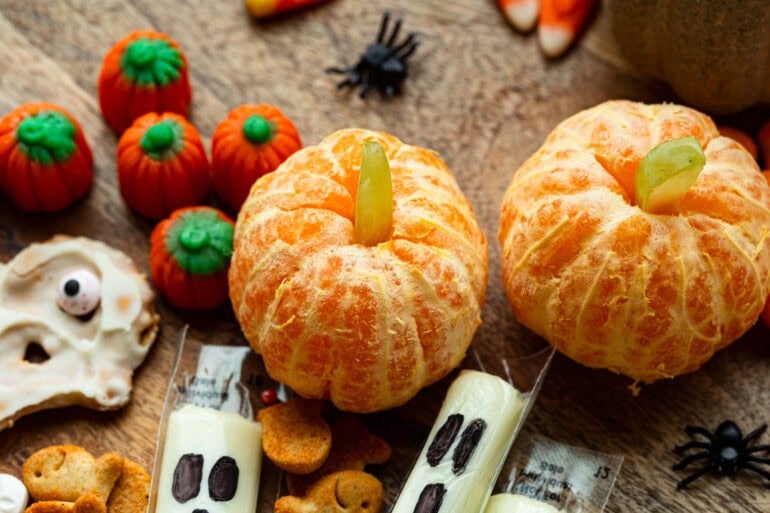 several halloween themed snacks scattered on a table including pumpkin tangerines, ghost cheese sticks, candy pumpkins