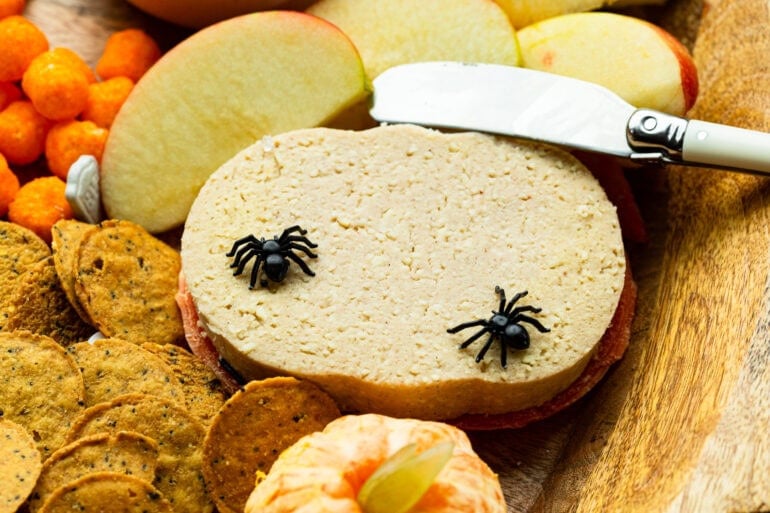 a chunk of pumpkin spice cheese sitting on a board with other halloween snacks with fake plastic spiders crawling on it