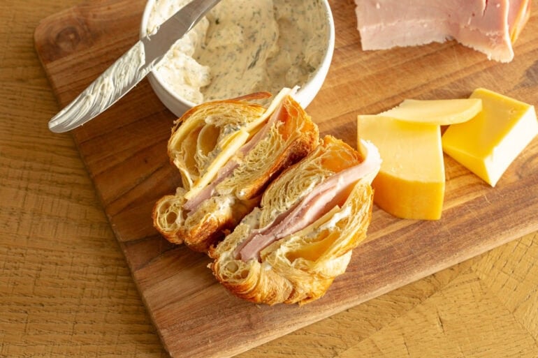 a croissant ham and cheese sandwich sliced in half with a block of gouda cheese sitting next to is and a small bowl of garlic herb spread