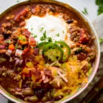 Award winning healthy turkey instant pot chili A metal bowl of ground turkey chili full of beans, red onions that have been chopped, chopped peppers , jalapeno slices on melted cheese next to a dollop of sour cream with a dash of cilantro and a grey napkin underneath