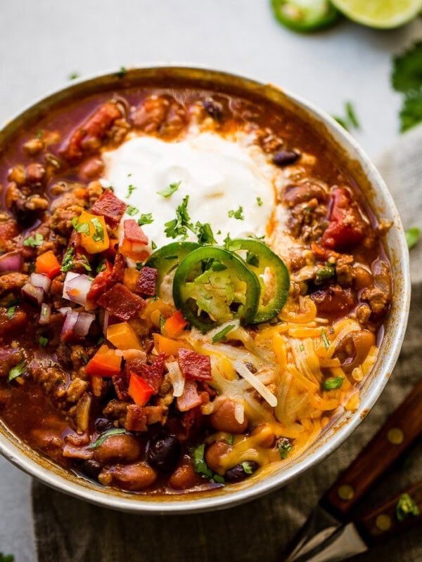 Award winning healthy turkey instant pot chili A metal bowl of ground turkey chili full of beans, red onions that have been chopped, chopped peppers , jalapeno slices on melted cheese next to a dollop of sour cream with a dash of cilantro and a grey napkin underneath