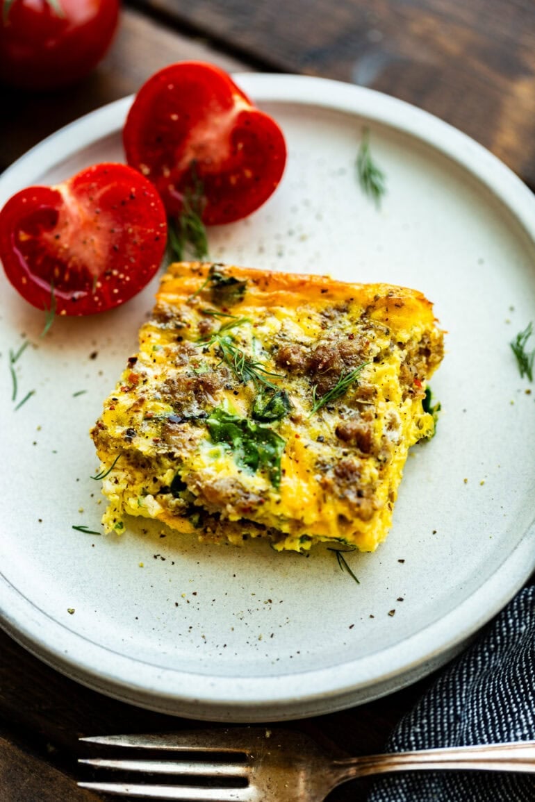 a serving of egg and sausage casserole with spinach on a white plate with a halved tomato sitting next to it