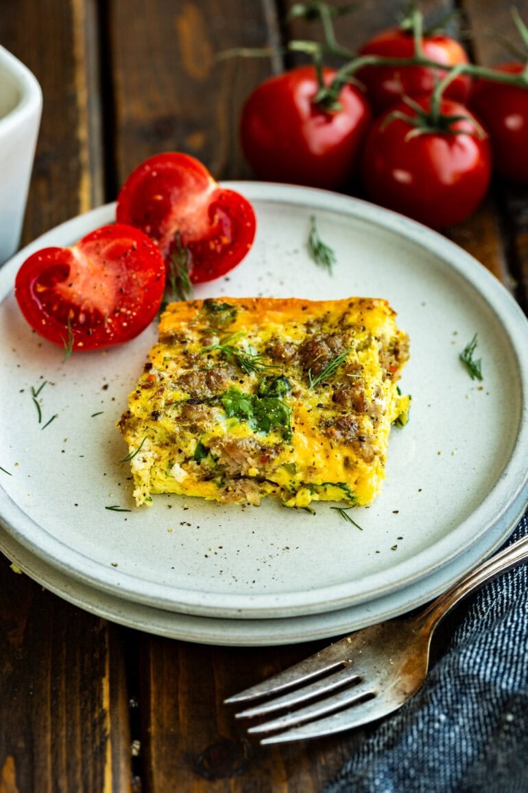 a piece of baked spinach sausage and egg casserole with a sliced tomato on the side