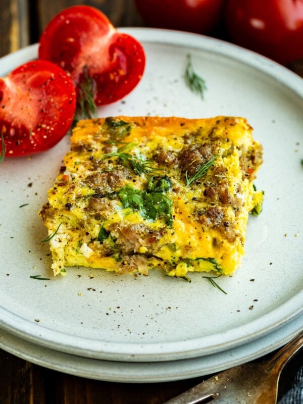 a serving of egg and sausage casserole with spinach on a white plate with a halved tomato sitting next to it