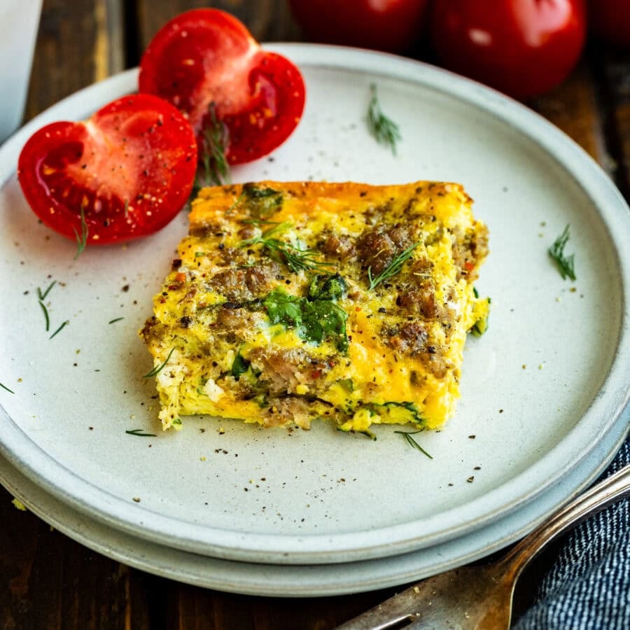 a serving of egg and sausage casserole with spinach on a white plate with a halved tomato sitting next to it
