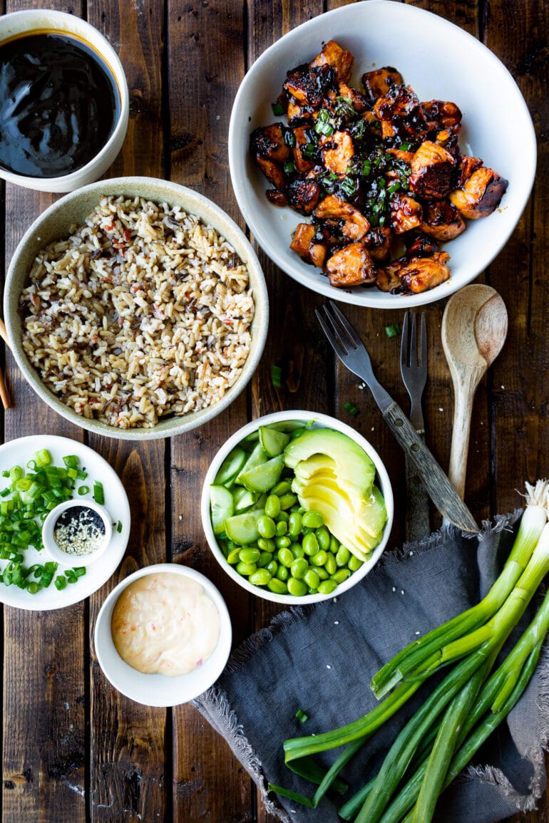 a photo of separate bowls small bowls with all the ingredients for teriyaki salmon bowls including teriyaki salmon bites, fluffy wild rice, sliced avocados, edamame beans, scallions and sesame seeds.