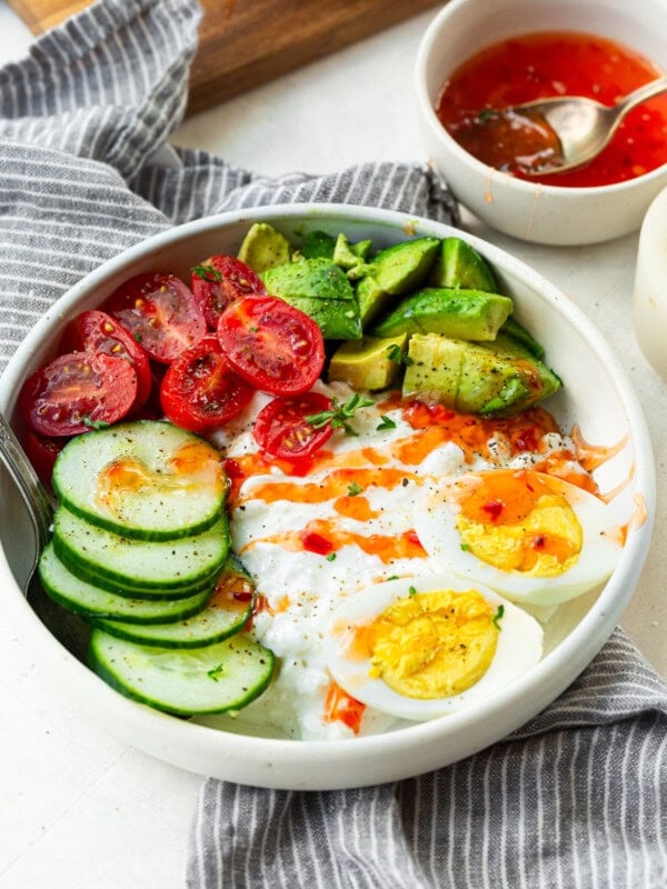 a photo of a large bowl full of cottage cheese, hard boiled eggs, sliced cucumber, cherry tomatoes and avocado topped with sweet chili sauce