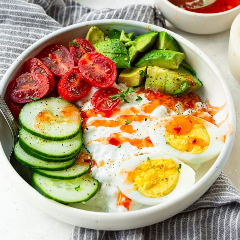 A photo of a large bowl filled with mozzarella cheese, hard-boiled eggs, cucumber slices, cherry tomatoes and avocado and topped with sweet chili sauce