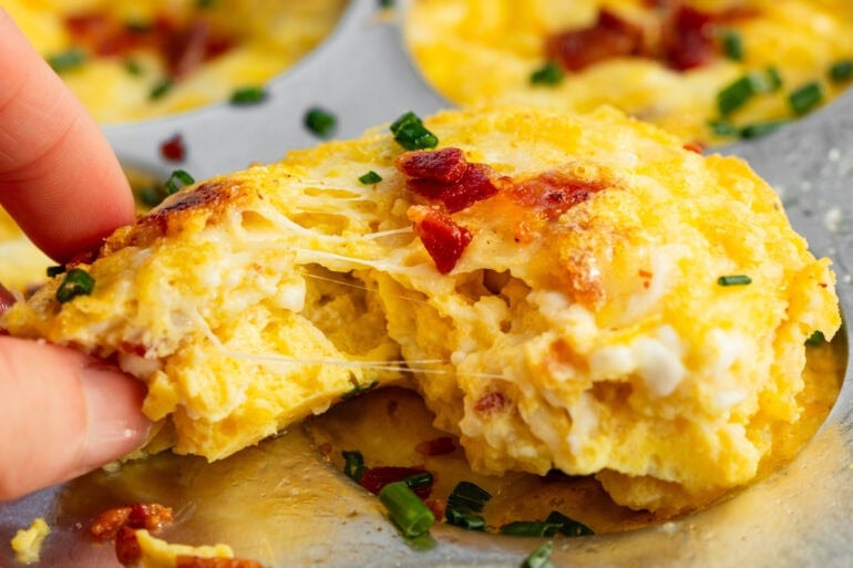 A close up of a muffin tin with cottage cheese egg bites garnished with bacon and chives. A hand is reaching and breaking apart an egg bite