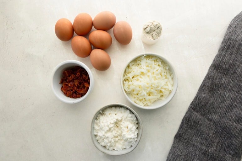 Ingredients for cottage cheese egg bites including 7 eggs, a scoop of protein powder, shredded cheese, bacon, and cottage cheese 