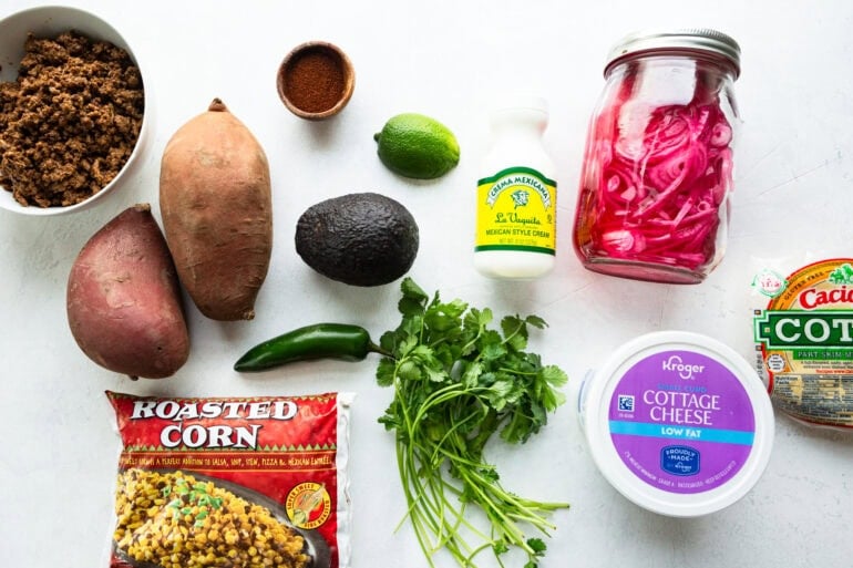 all the ingredients for street corn ground beef sweet potato bowl including sweet potatoes, mexican crema, cottage cheese, lime, avocado, jalapeno, cilantro, pickled red onions and corn