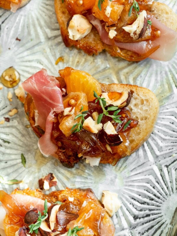 a photo taken over the top of a mandarin orange and prosciutto crostini topped with toasted hazelnuts and fresh time