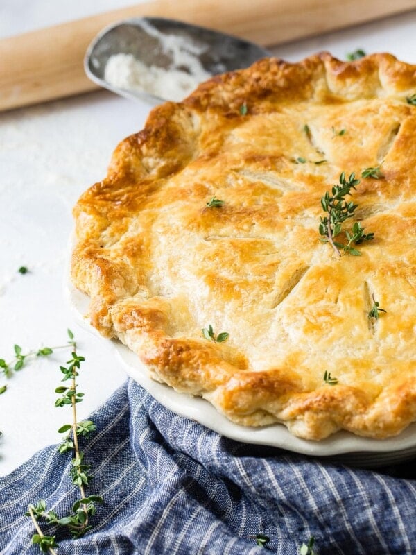 A chicken pot pie, baked to a perfect golden brown with a sprig of fresh thyme on top. The pie dish is sitting on a blue and white striped kitchen towel.